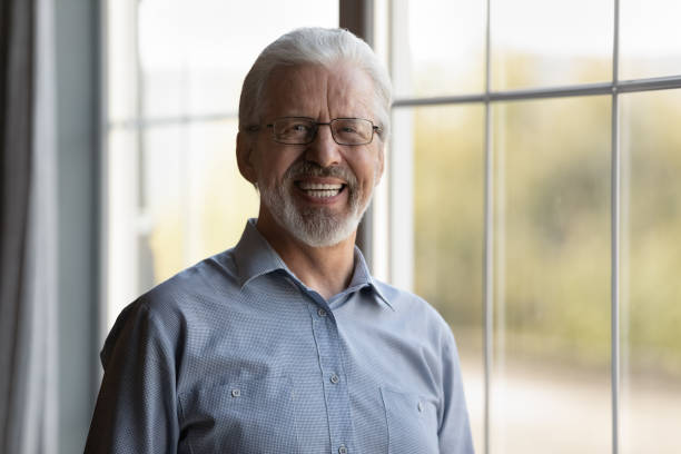 Headshot portrait of smiling mature Caucasian 60s man in glasses pose in own home apartment. Profile picture of happy middle-aged grey-haired 70s male or grandfather show optimism on maturity.